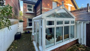 A conservatory with a glass roof, brick walls, and a small garden area with a white fence and a black trash bin. - Elm Villa (Wareham)