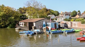 A riverside boat hire station with several boats docked, a small building with a sign, and houses in the background. - Elm Villa (Wareham)