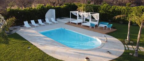 Una piscina rectangular con una terraza blanca alrededor, equipada con sillas de descanso y un área de estar cubierta.