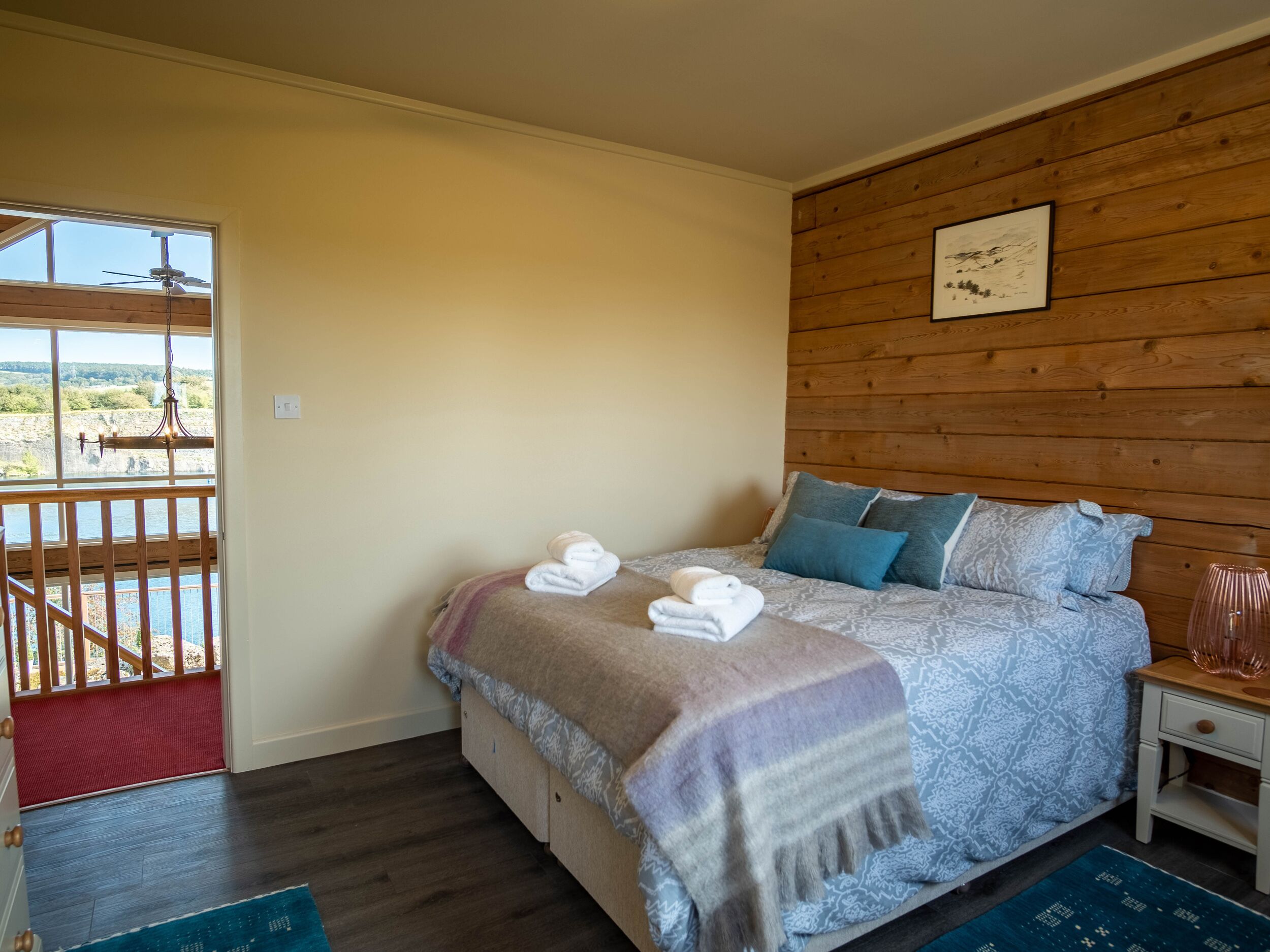 A bedroom with a bed, a nightstand, a wooden headboard, a window with a view, and a door leading to an outdoor area.