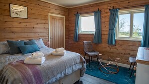 Um quarto aconchegante com parede de madeira, uma cama com almofadas, uma cadeira e uma mesa com tampo de vidro.