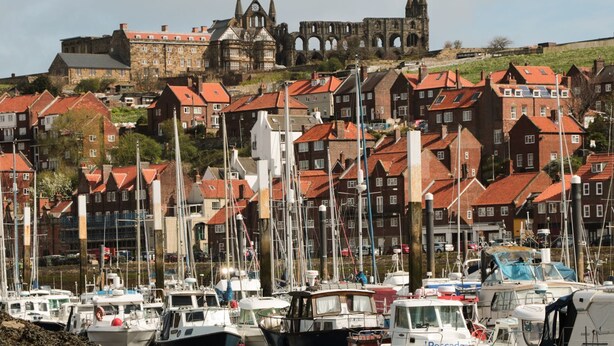 Ein Yachthafen mit vertäuten Booten, umgeben von Gebäuden mit roten Dächern und einer historischen Struktur im Hintergrund.