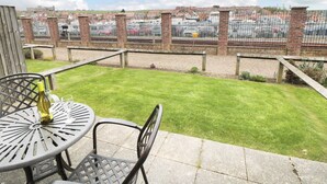 Eine Terrassenfläche mit Tisch und Stühlen, eine Rasenfläche und Blick auf einen Parkplatz mit Autos sowie ein Backsteingebäude im Hintergrund.