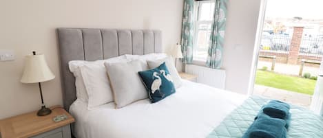 A bedroom with a grey upholstered headboard, white bedding, and a teal quilted bedspread. There is a wooden nightstand with a lamp, a window with patterned curtains, and a view of a building outside.