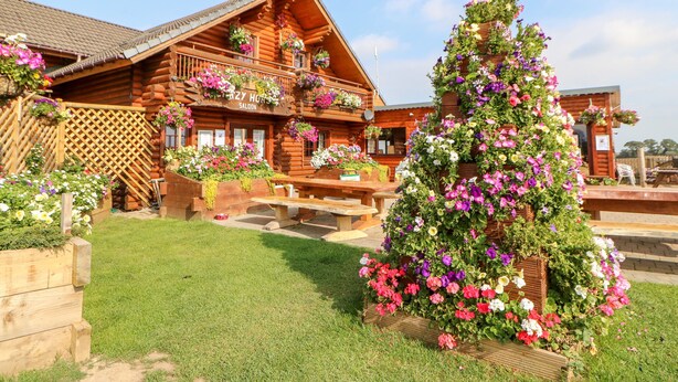 A wooden log cabin with a balcony adorned with flowers, a wooden deck, and a well-maintained lawn. - Jalan End (Morpeth)