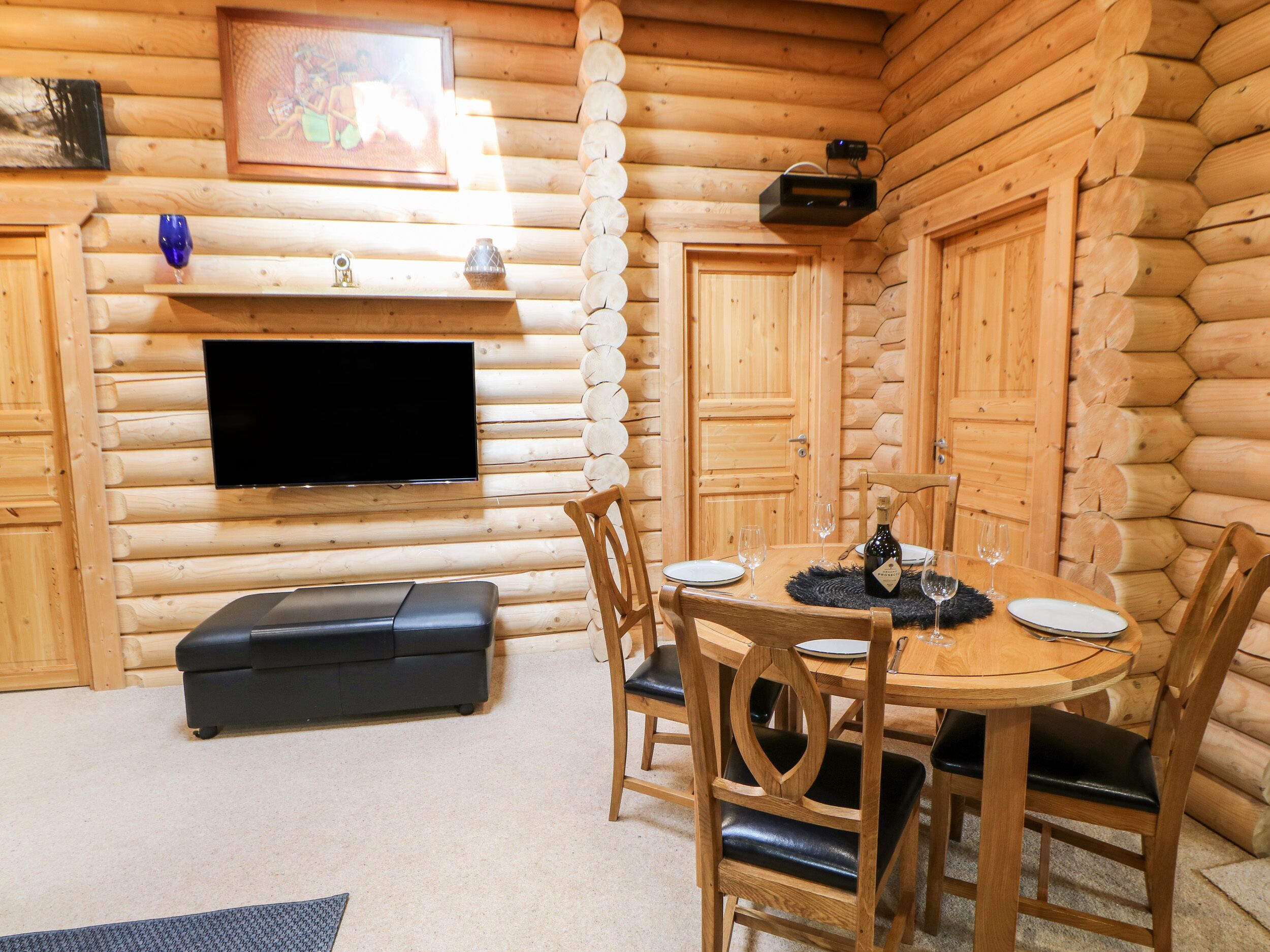 Interior de cabana de tronco com uma mesa de jantar redonda de madeira para quatro pessoas, rodeada por cadeiras de madeira com almofadas pretas. Uma televisão de tela plana está montada na parede acima da mesa, e uma lareira com mantelpiece é visível à esquerda.