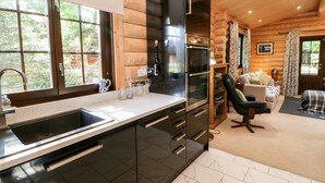 A modern kitchen with a black refrigerator, double oven, and stainless steel sink. There is a wooden dining table with chairs and a sofa in the living area. - Jalan End (Morpeth)