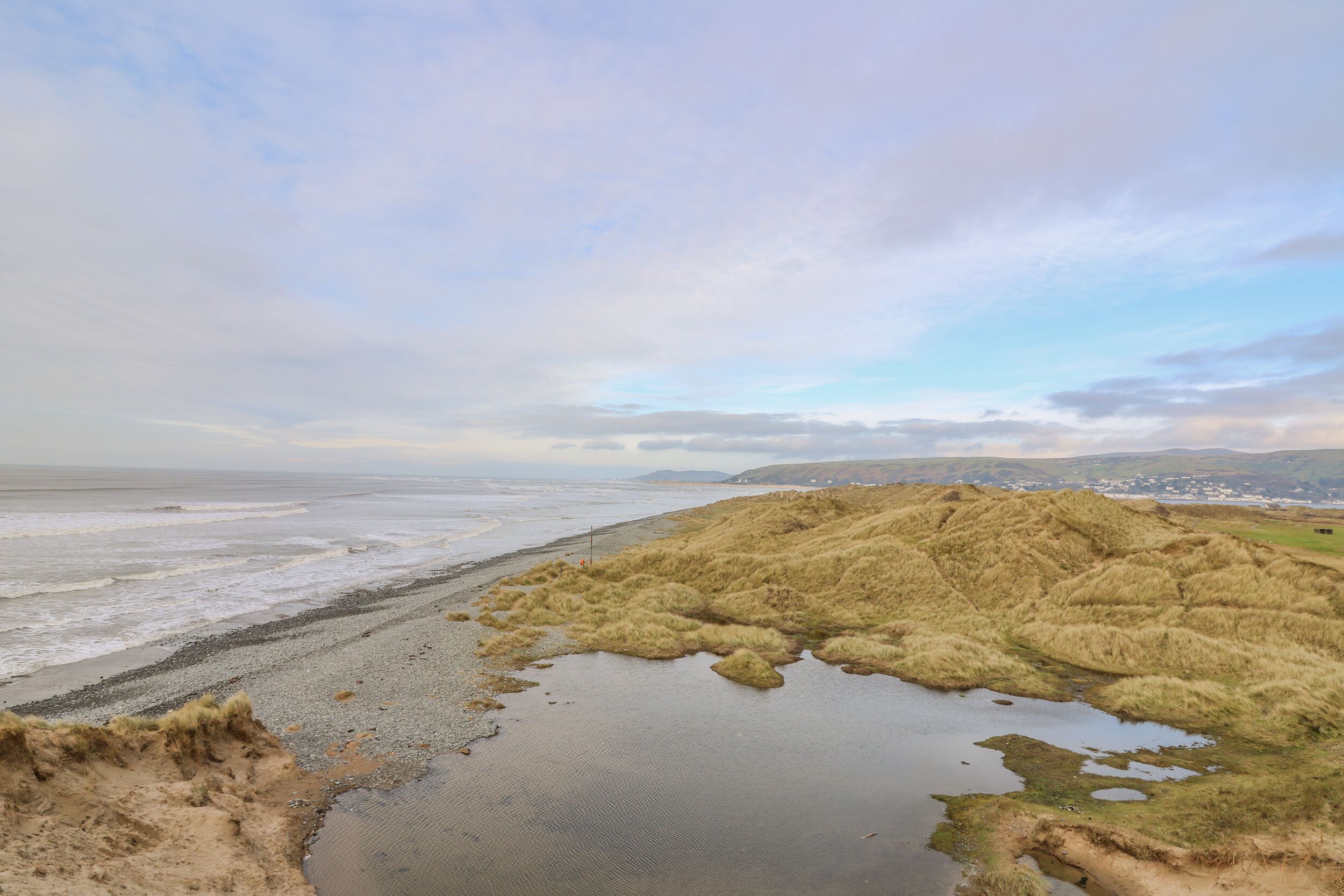 A coastal landscape with a beach, grassy dunes, and a small body of water.