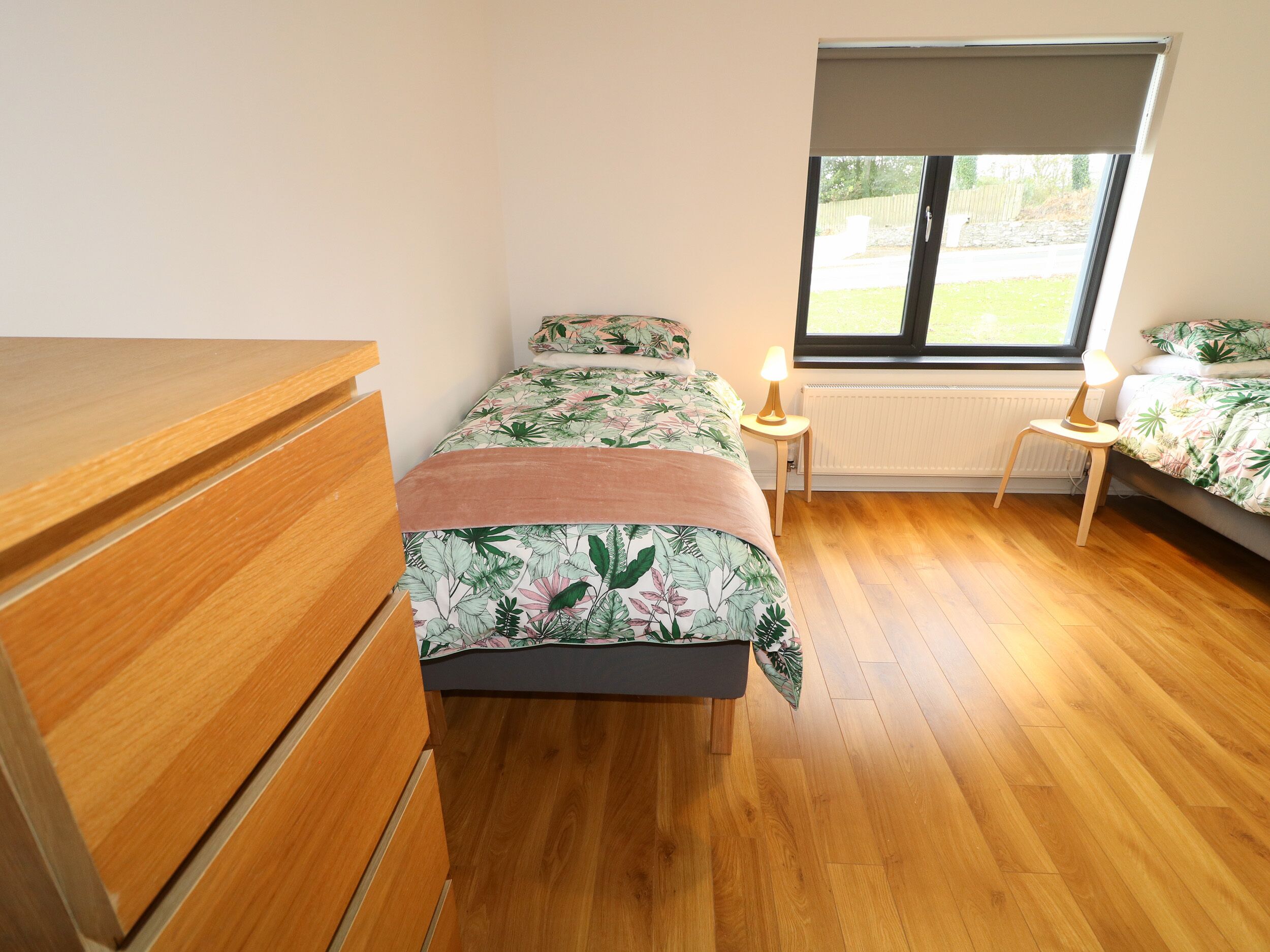 Quarto com cômoda de madeira, cama com roupa de cama floral, mesa de cabeceira com abajur e janela com vista para o exterior.