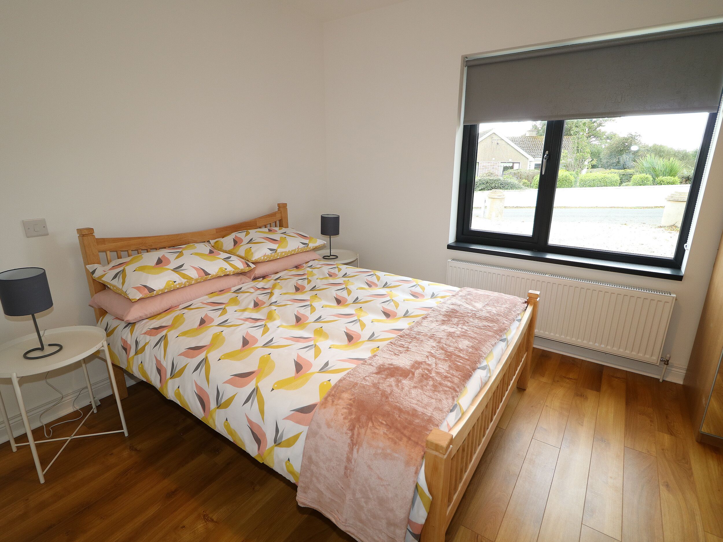 Un dormitorio con una cama de madera, una mesita de noche con lámpara, una ventana con vistas al exterior y un radiador debajo de la ventana.