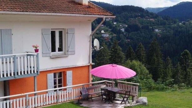 A two-story house with a balcony, a pink umbrella, and a view of a forested mountain range.