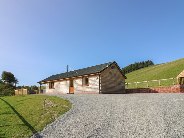 A small wooden cabin with a gravel driveway, surrounded by a grassy area and a wooden fence. - Ploony Hill Cabin (Knighton)