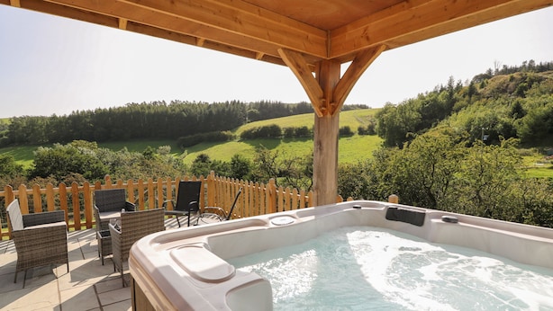 A hot tub with a wooden deck and chairs, overlooking a green landscape.