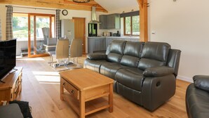 A spacious living room with a leather sofa, wooden coffee table, and a TV. A kitchen with modern appliances and a dining area with chairs are visible in the background.