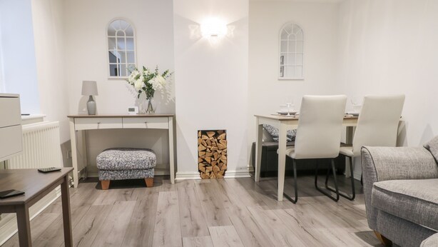 A modern living room with a wooden floor, a white sofa, a dining table with chairs, a small desk with a lamp, and a fireplace. - Swallow Cottage (Herne Bay)