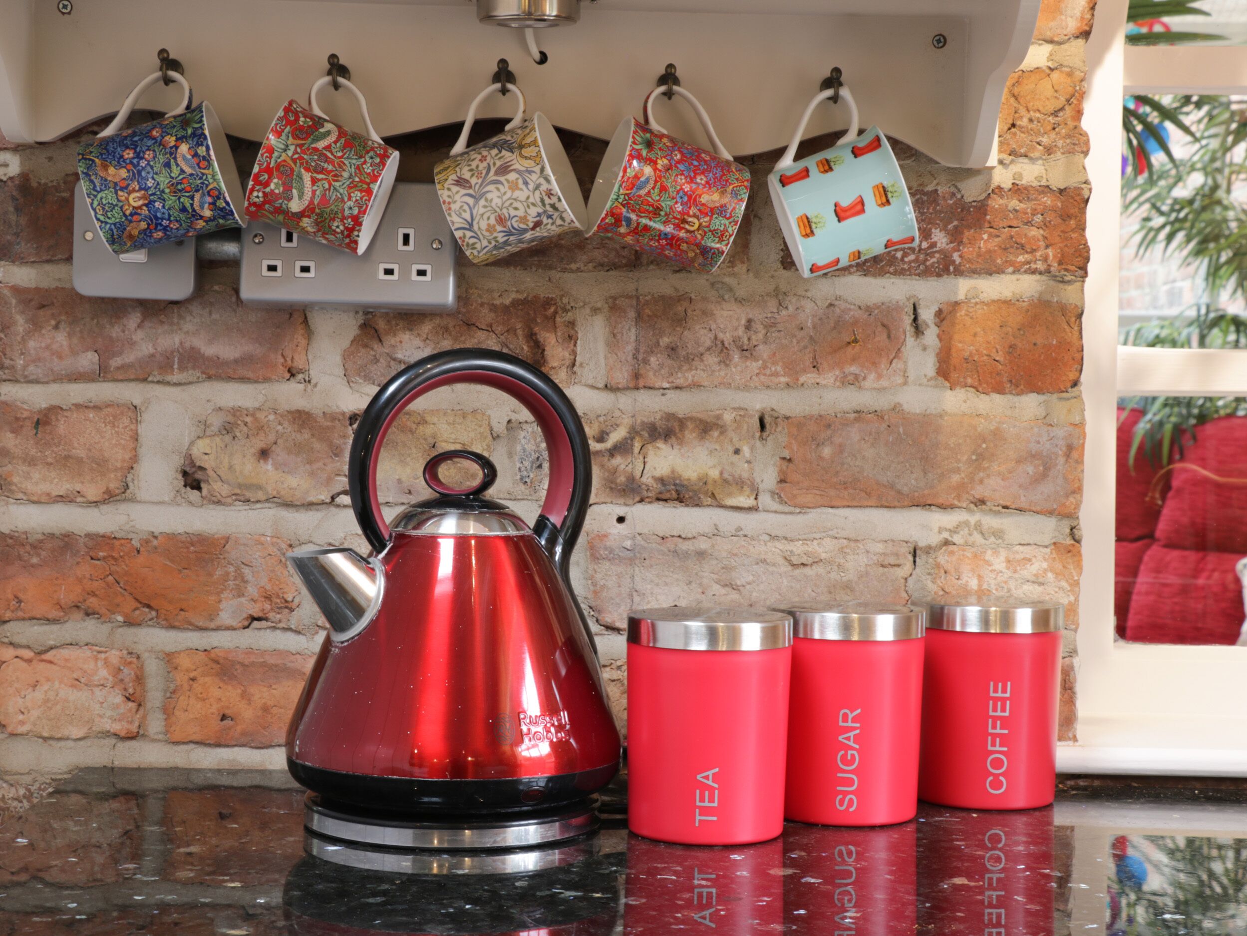 A kitchen with a red kettle, colorful mugs, and red containers labeled 'TEA', 'SUGAR', and 'COFFEE'.