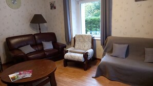 A living room with a brown leather sofa, a patterned armchair, and a grey sofa. There is a wooden coffee table with magazines and a lamp.