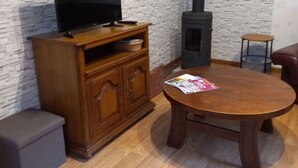 A living room with a television on a wooden cabinet, a round wooden table with a magazine, a small round stool, and a wood-burning stove.