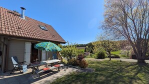 A house with a tiled roof, a covered patio with a table and chairs, and a lawn with trees. - Cozy Chalet near Plombières-les-Bains - Ideal for Couples, Nature Trails, and Relaxing Spa Retreat (Le Val-d'Ajol)