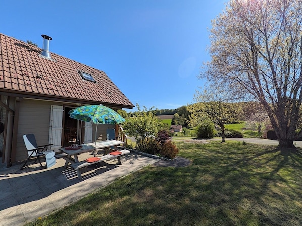 A house with a tiled roof, a covered patio with a table and chairs, and a lawn with trees.