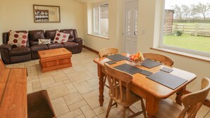 A living room with a brown leather sofa, a wooden dining table with chairs, a coffee table, and a large window with a view of a garden. - Bron Gadair (Llanfairpwllgwyngyll)
