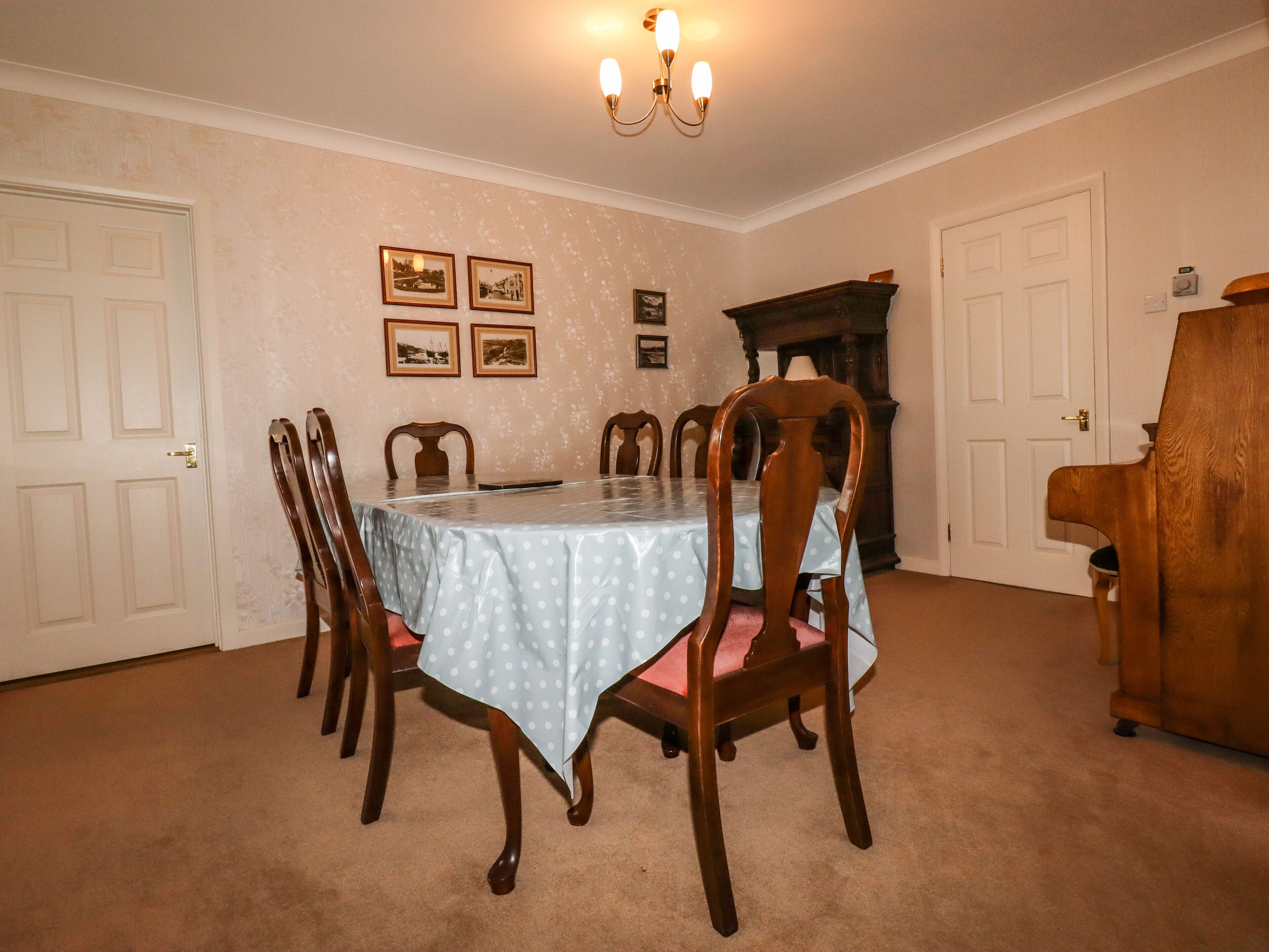 Uma sala de jantar com uma mesa posta para seis pessoas, rodeada por cadeiras de madeira. Há quadros nas paredes, uma lareira e um piano ao lado.