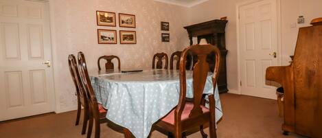 Uma sala de jantar com uma mesa posta para seis pessoas, rodeada por cadeiras de madeira. Há quadros nas paredes, uma lareira e um piano ao lado.