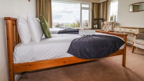 Une chambre d’hôtel avec un lit en bois, un bureau et une chaise. Il y a une grande fenêtre avec des rideaux, et la vue donne sur la ville.