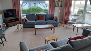 Un salon avec un canapé gris, une table basse, un téléviseur posé sur un meuble et une vue sur une maison à travers de grandes fenêtres.