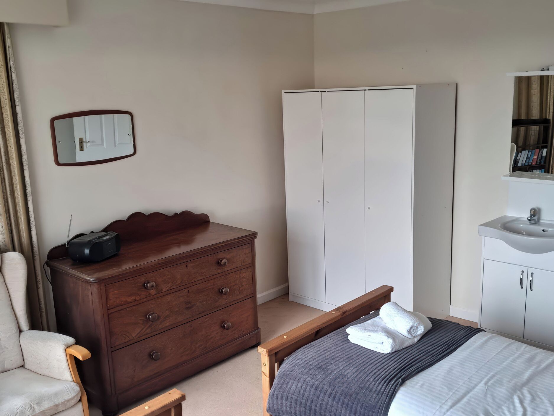 A bedroom with a bed, a wooden dresser, a wardrobe, a bedside table, a mirror, and a bathroom with a sink.