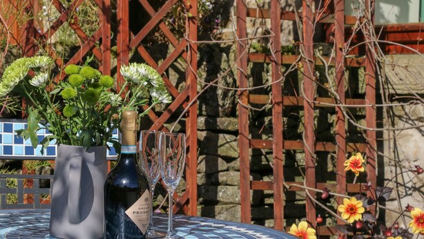 A round table with a bottle of organic prosecco and two glasses, surrounded by a mosaic table top and a wooden fence with flowers. - The Old Cart House (Hope Valley)
