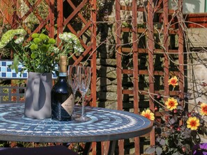 A round table with a bottle of organic prosecco and two glasses, surrounded by a mosaic table top and a wooden fence with flowers. - The Old Cart House (Hope Valley)