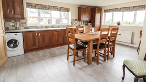 A kitchen with wooden cabinets, a washing machine, a dining table with chairs, and large windows. - Sandmartins (Chester)