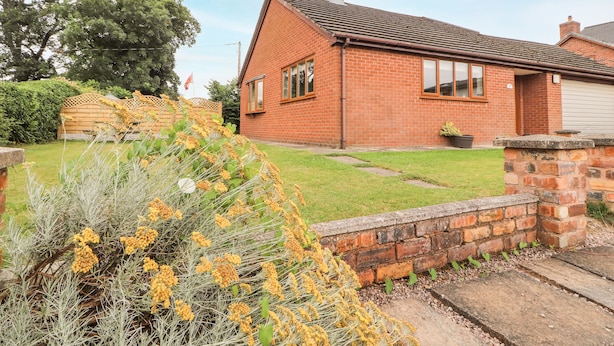 A brick house with a tiled roof, a front yard with grass and plants, and a stone wall. - Sandmartins (Chester)