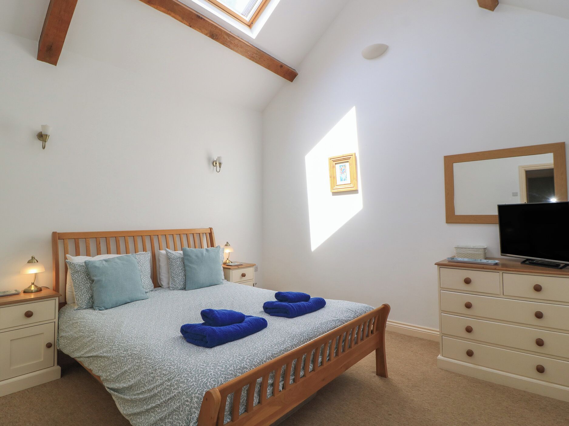 A bedroom with a wooden bed, bedside tables, a dresser, a TV, and a skylight.