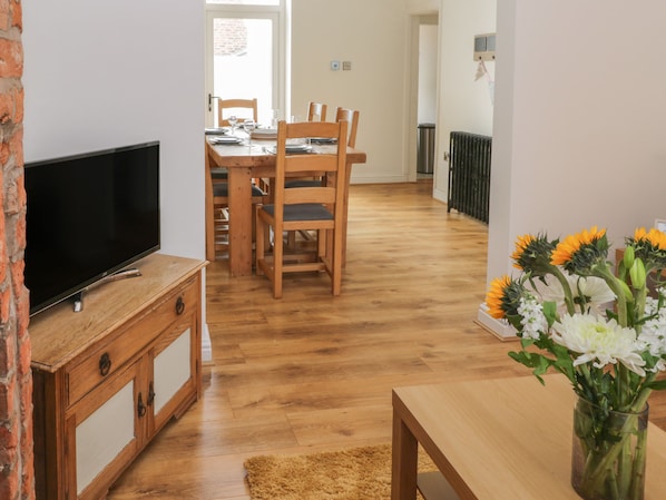 A room with a TV on a wooden cabinet, a dining table with chairs, and a vase with flowers. - Laridae (Whitby)
