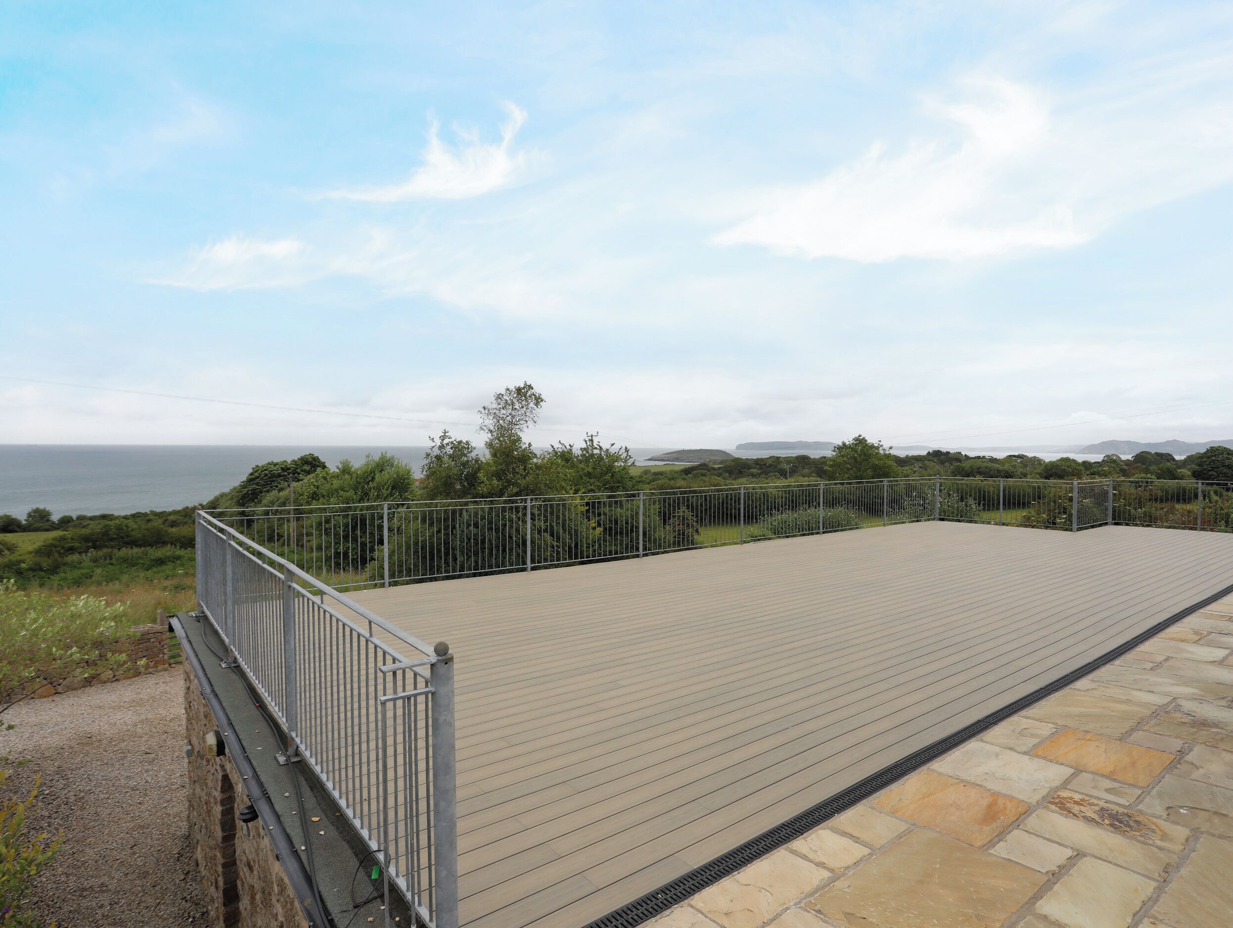 A flat roof terrace with a metal railing, overlooking a coastal landscape with greenery and distant hills.