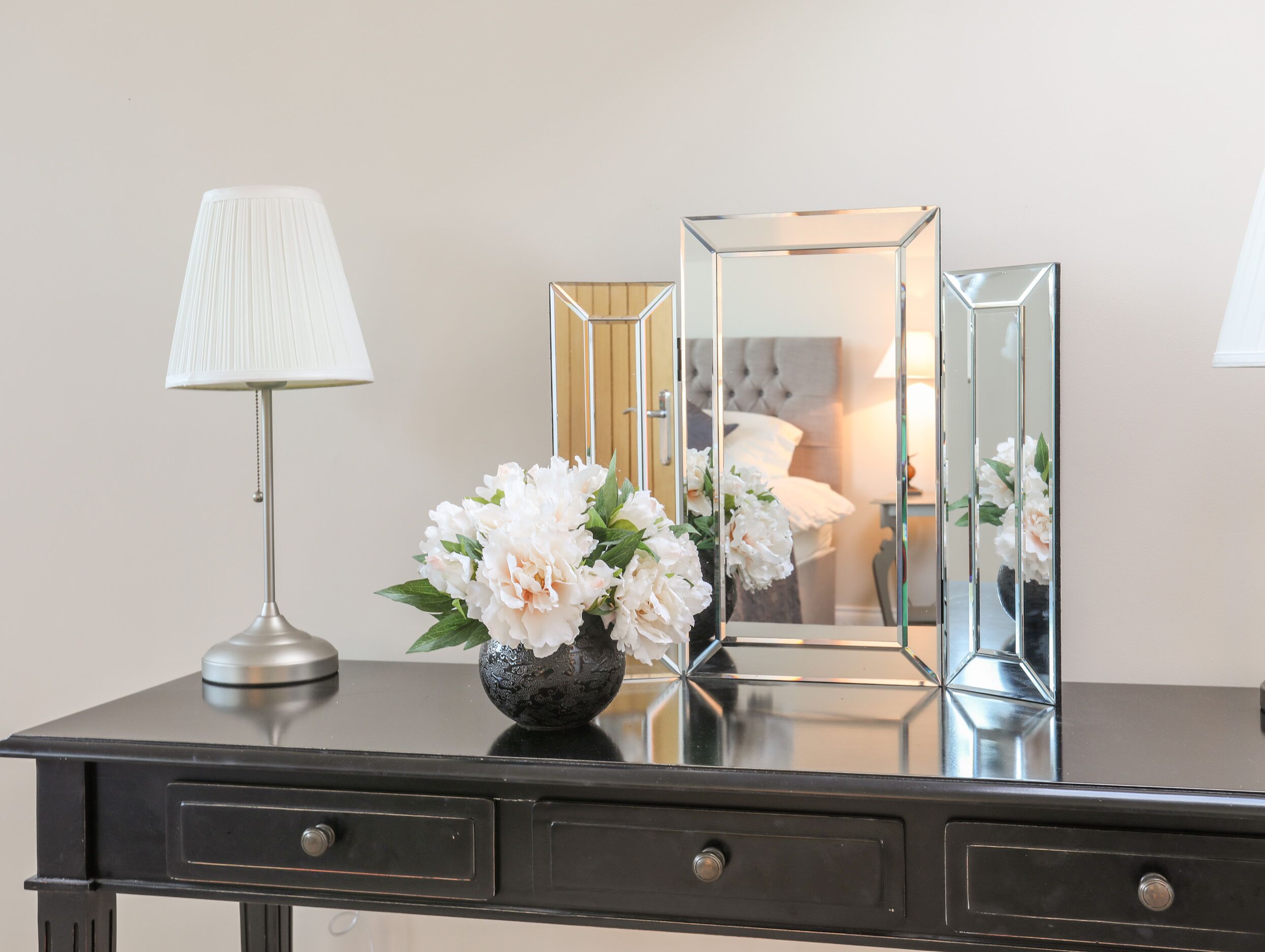 A black desk with a lamp, a vase with flowers, and three mirrors.