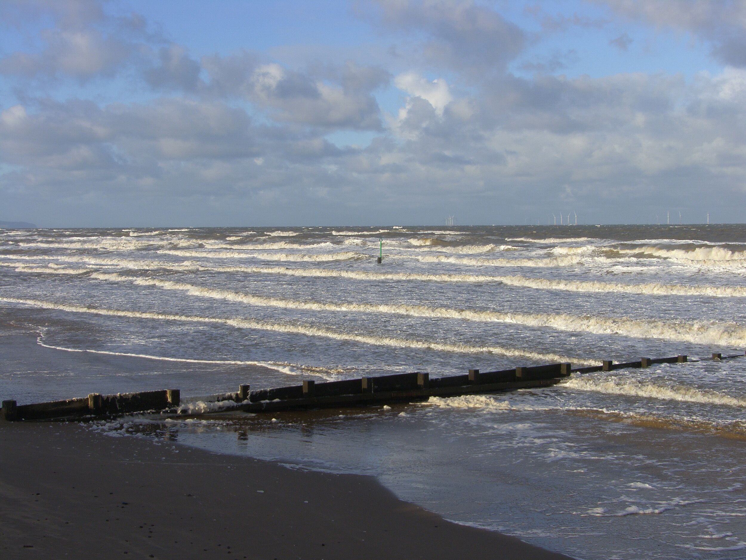 Ein Strand mit brechenden Wellen, einer Holzsteg und Windkraftanlagen in der Ferne.
