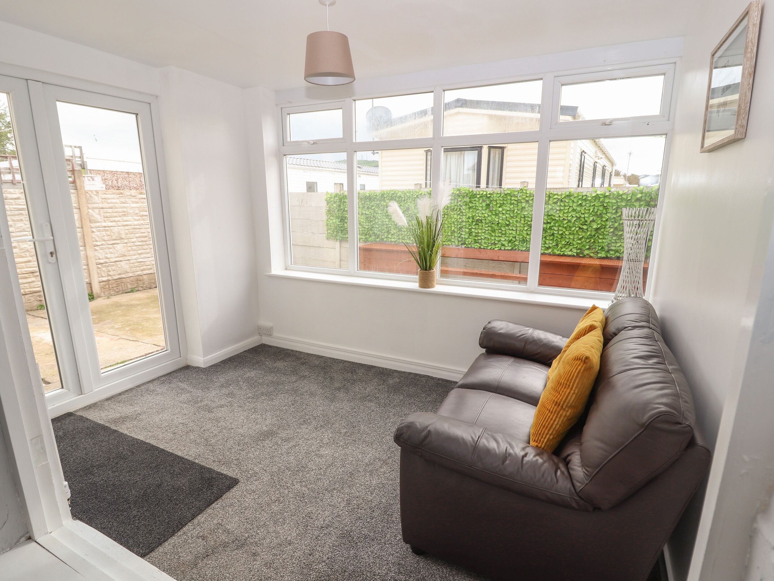 A room with a grey carpet, a brown leather sofa with a mustard throw pillow, a window with a view of a green hedge and a brick wall, and a hanging light fixture.