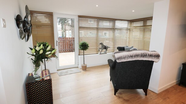 A modern living room with a dark grey sofa, a wooden floor, a potted plant, and a window with blinds. - Swale Cottage (Chathill)