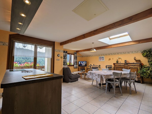 Un salon chaleureux avec un coin repas, des poutres en bois, un téléviseur et une grande fenêtre donnant sur un paysage verdoyant.