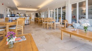 A spacious dining area with wooden tables and chairs, a kitchen in the background, and large windows offering views of the outdoors. - 17 Burgh Island Causeway (Kingsbridge)