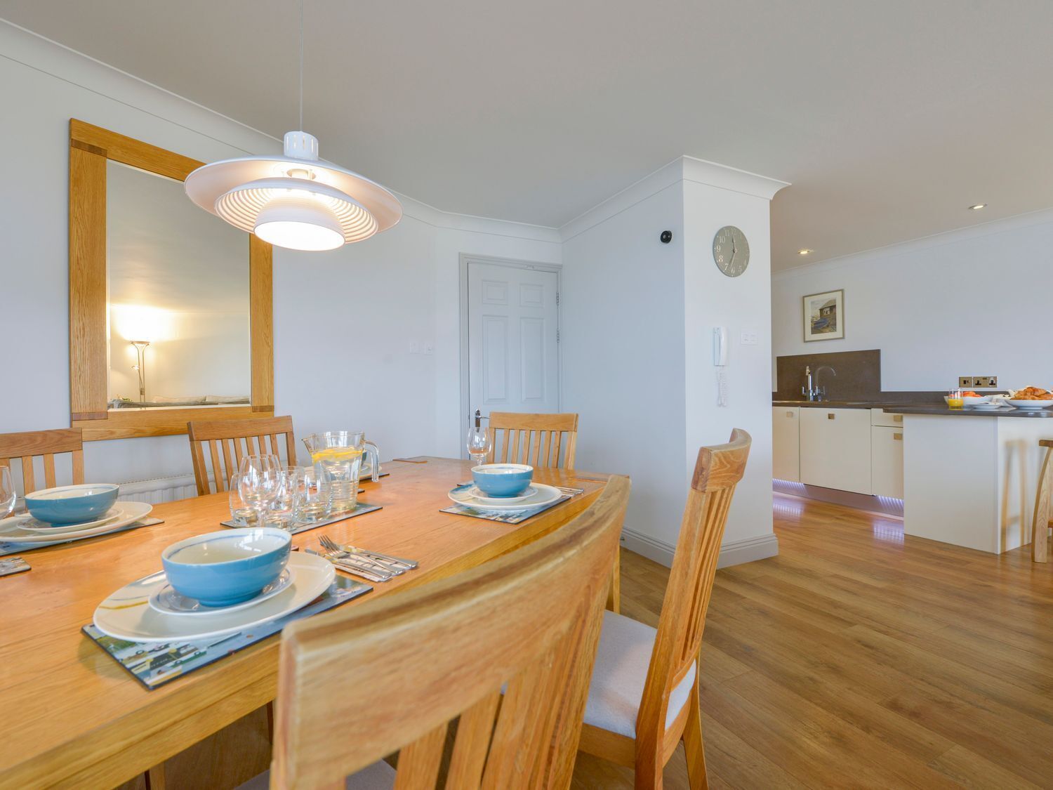 A dining area with a wooden table set for four, a large mirror, and a kitchen in the background.