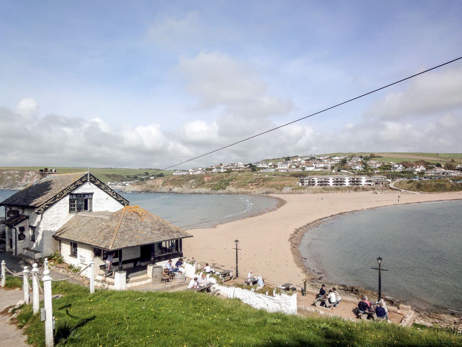 A coastal scene with a beach, a small white building with a thatched roof, and a view of houses in the distance.