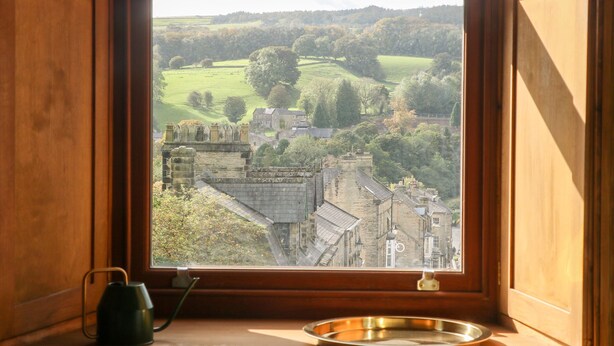 Vista a través de una ventana de un edificio histórico con arquitectura de piedra, un paisaje verde y un cielo despejado.