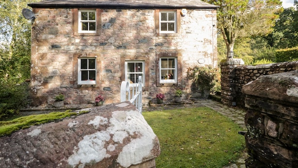 A stone house with a central door, two windows on each side, and a small garden area with a stone wall and a pathway. - Fell Foot (Holmrook)