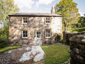A stone house with a central door, two windows on each side, and a small garden area with a stone wall and a pathway.