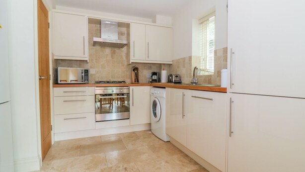 A modern kitchen with white cabinets, a built-in oven, a microwave, a washing machine, and a window providing natural light. - 26 Burtons Mill (Norwich)