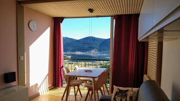Una habitación con una mesa de comedor y sillas, un sofá con un cojín de lobo, un reloj en la pared y vistas a montañas a través de grandes ventanales.
